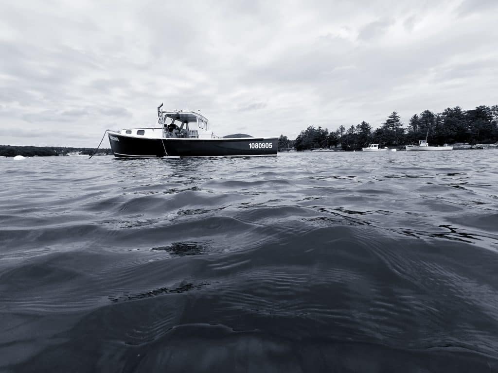 A striking black and white photograph capturing the tranquil scene of a lobster boat moored in Blue Hill Bay, Blue Hill, Maine. The calm waters and serene setting reflect the quintessential coastal charm of the area, with the lobster boat, marked with the number 1080905, taking center stage against a backdrop of lush, forested shoreline. The monochromatic tones enhance the timeless quality of the image, showcasing the boat's sleek lines and the gentle ripples on the water.