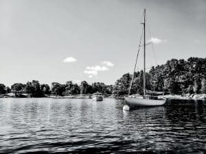 This black and white photograph captures the serene beauty of Blue Hill Bay, located in Blue Hill, Maine. The composition features a tranquil scene with sailboats moored on calm waters, set against a backdrop of lush trees and a clear sky. The reflective quality of the water adds a sense of depth and contrast to the image, emphasizing the natural elegance and tranquility of the coastal setting.