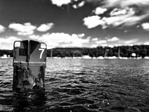 This dramatic black and white photograph highlights a channel marker, number 7, standing resolutely in the waters of Blue Hill Bay, Maine. The stark contrast in the image emphasizes the rugged texture of the buoy and the shimmering surface of the water, while the cloud-filled sky provides a dynamic backdrop. The focused composition on the buoy, set against the expansive view of the bay and distant sailing boats, captures a moment of stillness and the enduring role of such markers in maritime navigation.