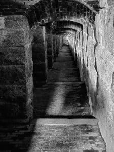 This photograph captures a historic hallway within Fort Knox in Bucksport, Maine. Fort Knox is a significant military structure from the mid-19th century, renowned for its well-preserved granite architecture. The image highlights the intricate brick and stonework, with the long, shadowy corridor leading to a distant light, emphasizing the fort's imposing and mysterious atmosphere. The hallway, with its repetitive arches and stone textures, reflects the craftsmanship and strategic design of the era, offering a glimpse into America's military past.