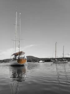 This photograph, titled "Mountain View from the Bay," captures the serene beauty of a sailboat anchored in a calm bay, with a mountainous backdrop adding depth and grandeur to the scene. The color pop effect emphasizes the bright orange hues of the boat’s details, contrasting against the grayscale tones of the surrounding water and sky. The reflection of the boat on the still waters enhances the tranquility of the moment, evoking a sense of peace and connection to nature. This image beautifully highlights the serene coastal landscapes of Downeast Maine.