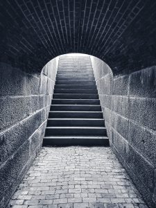 This photograph showcases a stairway at Fort Knox, a historic military fortification located in Bucksport, Maine. The image captures the stone and brick architecture of the fort, with the stairs leading upward from a dark, tunnel-like passageway toward a brighter, light-filled exit. Fort Knox, constructed between 1844 and 1869, was built to protect the Penobscot River and the surrounding region from potential British naval threats. The black and white composition emphasizes the texture of the weathered stones and bricks, creating a sense of mystery and historical depth as the viewer's eye is drawn up the stairs, symbolizing a journey from darkness into light.