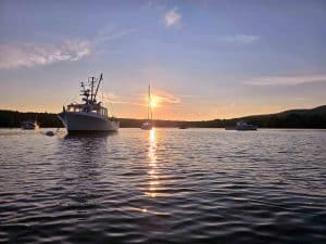 This evocative photograph captures the serene beauty of a sunset over Blue Hill Bay in Blue Hill, Maine. The tranquil waters of the bay reflect the warm evening light, illuminating a cluster of lobster boats and sailboats. The scenic backdrop of Blue Hill adds to the picturesque coastal landscape, epitomizing the peaceful maritime charm of Downeast Maine. The soft colors and calm atmosphere invite viewers to experience a moment of tranquility and appreciation for the natural beauty of the region.