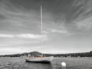 This black and white photograph captures a sailboat moored in Blue Hill Bay, Maine, with Blue Hill Mountain looming in the background. The choice to render the image in black and white focuses attention on the contrasts and textures of the water and sky, while the subtle hues of the boat highlight its serene presence against the vast landscape. This artistic approach underscores the tranquility and timeless beauty of Maine's coastal scenery.