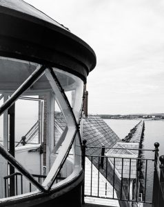 The Rockland Breakwater Lighthouse in Rockland, Maine is a stunning sight, especially during the annual lighthouse opening event in September. To reach the lighthouse, visitors must traverse a man-made rocky breakwater that stretches nearly a mile. It was during one of these treks that I stumbled upon this breathtaking scene. As I climbed to the top of the lighthouse, I noticed a boat in the distance making its way towards us. Without a camera stand, I leaned over the rail and steadied my camera loaded with 35mm slide film. The ship soon came into view and crossed in front of the lighthouse, making for a truly unforgettable moment.