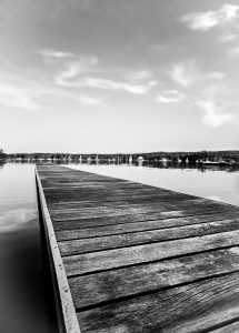 This black and white photograph captures a peaceful dock extending into the calm waters at a boatyard in Blue Hill, Maine. The perspective of the image draws the viewer's eye down the length of the wooden planks, leading toward the distant shoreline dotted with boats and surrounded by lush forests. The quiet stillness of the scene, coupled with the minimalistic composition, evokes a sense of tranquility and timelessness. The gentle ripples on the water and the soft clouds in the sky add subtle texture and depth to this serene coastal landscape.