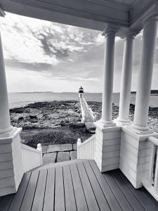 This photograph captures a breathtaking view from the porch of the lighthouse keeper's house at Marshall Point Lighthouse, located in Port Clyde, Maine. The image is framed by the white columns of the porch, leading the viewer's eye down the wooden walkway toward the iconic lighthouse, which stands resolute at the end of a rocky shoreline. The lighthouse, famous for its appearance in the movie Forrest Gump, is a symbol of maritime heritage, offering guidance to ships navigating the rugged Maine coast. The black and white treatment of the photograph enhances the timelessness of the scene, emphasizing the contrast between the sturdy architecture and the natural beauty of the surrounding landscape.