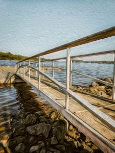 This artistic photograph captures the gangway leading down to the dock in Blue Hill Bay, located in Blue Hill, Maine. The image is rendered with a painterly effect, enhancing the textures and colors of the wooden gangway, the surrounding rocks, and the calm waters of the bay. The perspective of the gangway creates a sense of depth, inviting the viewer to step into the scene and experience the tranquility of the coastal setting.