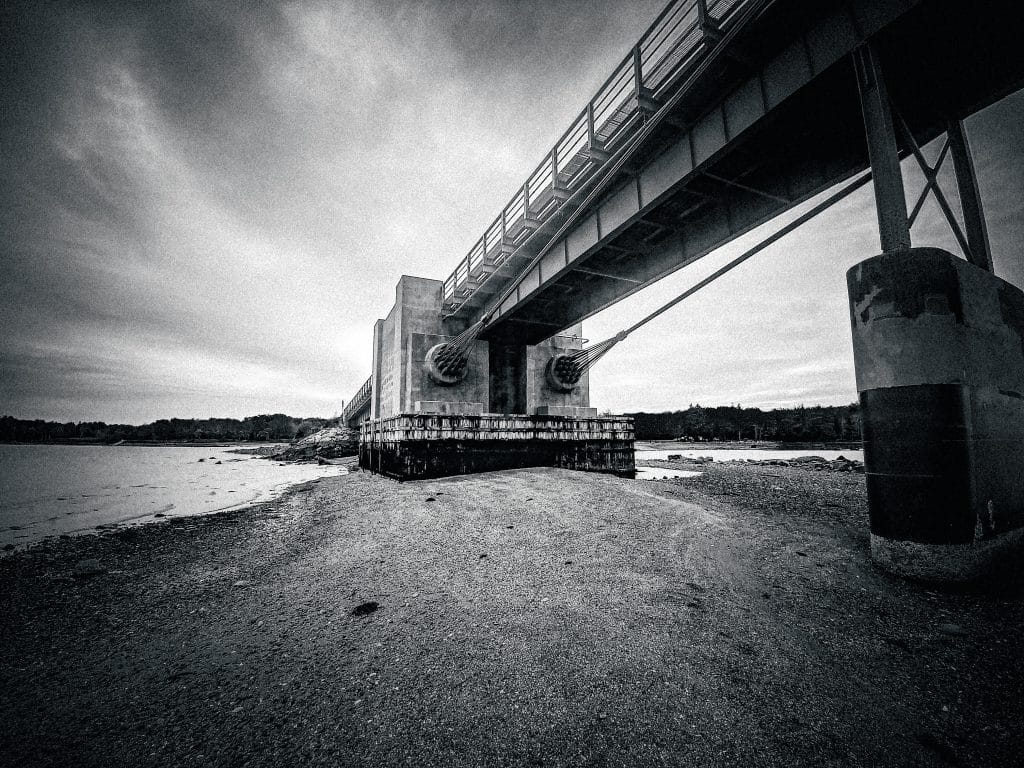 The image depicts the Deer Isle Bridge, a striking and iconic structure located in Downeast Maine. The photograph is taken from beneath the bridge, showcasing its massive support columns and cables, which stretch out dramatically toward the horizon. The overcast sky and the grayscale tones give the image a moody, atmospheric quality, emphasizing the rugged beauty of the Maine coastline. The composition leads the viewer’s eye along the bridge, creating a sense of depth and scale, while the calm waters and distant shoreline provide a serene contrast to the industrial strength of the bridge.