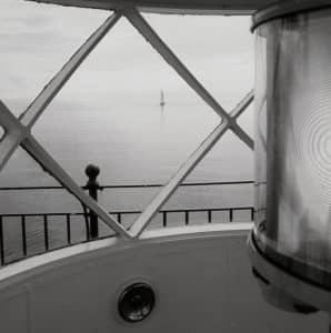 "View from the Lighthouse" captures a serene black-and-white scene of a sailboat in the distance, framed by the window of a lighthouse lantern room. A timeless and peaceful coastal moment.