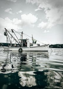 This photograph captures a working boat anchored in Blue Hill Bay, Blue Hill, Maine. The boat stands out against the backdrop of a dramatic sky and tranquil waters, highlighting the daily maritime activities that are a staple in this region. The image emphasizes the serene yet industrious nature of coastal life, with reflections on the water enhancing the sense of calm and the boat symbolizing the hard-working spirit of the local community.