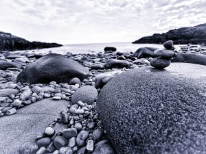 This photograph captures the rugged beauty of Little Hunters Beach in Acadia National Park, Maine. The image showcases a serene coastal scene with smooth, weathered stones of varying sizes spread across the shoreline. A small cairn, delicately balanced atop one of the larger rocks, adds a human touch to this natural setting. The perspective is low to the ground, emphasizing the texture and details of the stones, while the ocean and rocky cliffs in the background hint at the expansive landscape beyond. The soft, diffused light and muted colors create a calm and contemplative mood, perfectly encapsulating the raw beauty of Acadia's coastline.