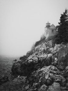 Black and white photograph of Bass Harbor Lighthouse shrouded in fog, perched on a cliff above the ocean, captured by Blaine Stoner.