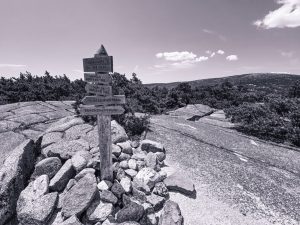 Monochrome photo of a wooden trail marker at the summit of Champlain Mountain in Acadia National Park, with Cadillac Mountain visible in the background.