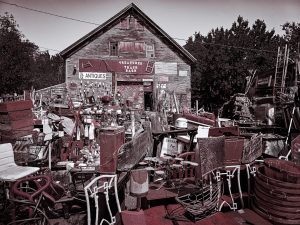 Photograph of an overflowing antique store in Searsport, Maine, with vintage signs, tools, and assorted objects stacked around a rustic barn-like building.
