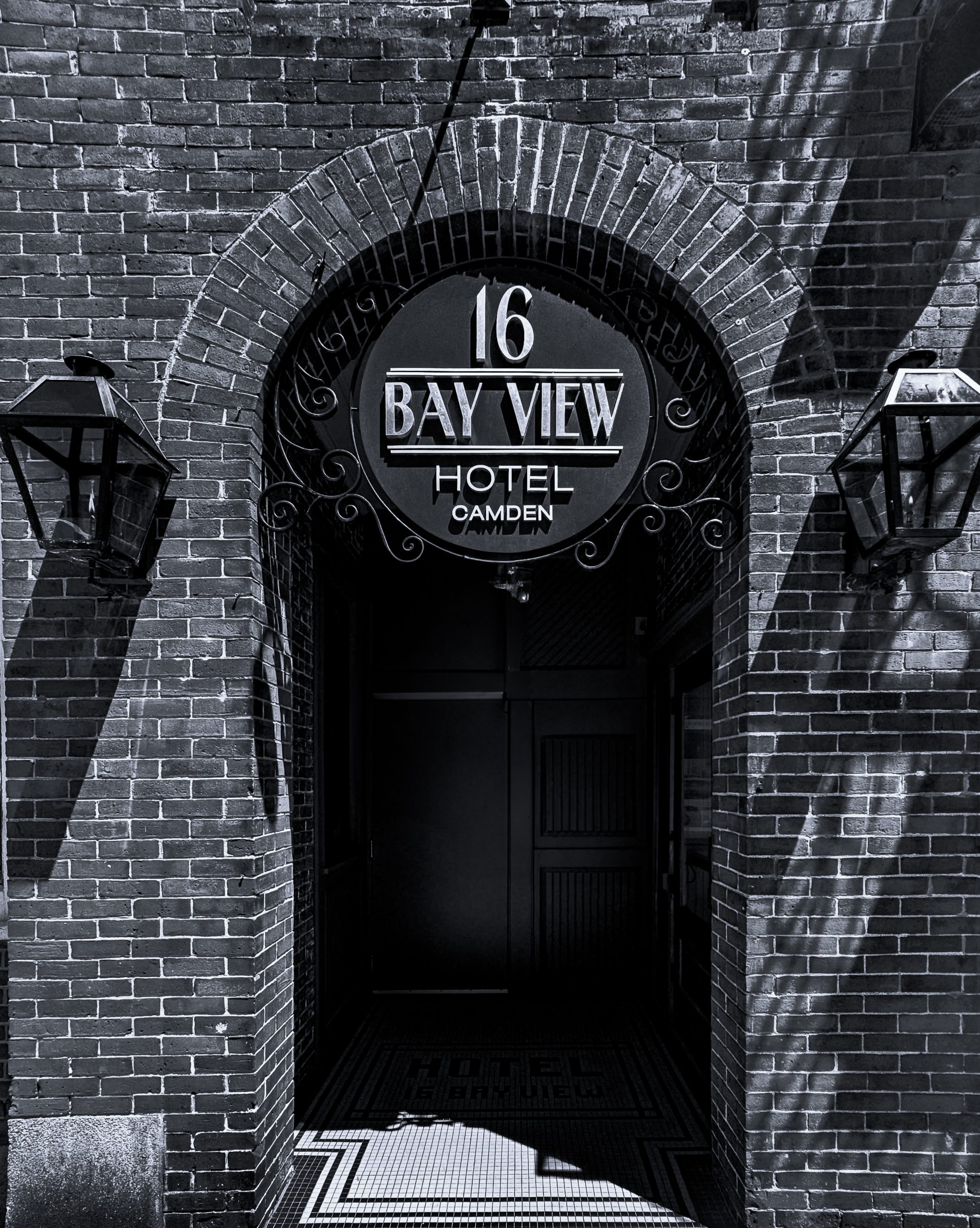 In this bold monochromatic portrait of Camden’s Bay View Hotel, Blaine Stoner captures a striking interplay of brick, shadow, and typography. The arched brick entryway frames the hotel’s art deco-inspired signage with precision, while twin lanterns cast geometric shadows across the historic facade. Sunlight filters through unseen trees, painting the red brick walls with rhythmic patterns of light and contrast. The image stands at the crossroads of street photography and architectural study—a still moment of balance between craftsmanship and contemporary design. With its sharp textures and moody lighting, “Bay View Shadows” evokes the quiet dignity of Camden’s coastal charm, while nodding to the elegance found in the simplest corners of its downtown architecture.