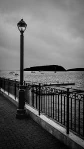 A lamppost and iron railing frame the view of Frenchman Bay in Bar Harbor, Maine, with boats scattered across the water under a moody sky.