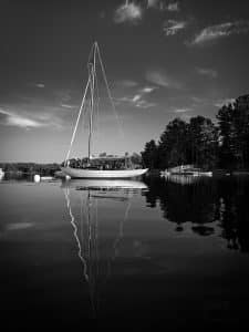 A lone sailboat rests peacefully in Blue Hill Bay, Maine, its mast perfectly mirrored in the still waters under a soft sky.