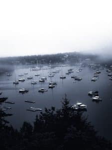 Foggy morning view of Northeast Harbor from Asticou Gardens in Maine, with boats anchored on calm water and mist over the shoreline.