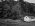 A high-contrast black and white photograph of a small, white weathered wooden boathouse with rusted boat rails extending down a pebbled shoreline, set against a dense forest in Belfast, Maine.