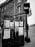 A black-and-white low-angle photograph of a street corner in downtown Belfast, Maine, featuring a dark kiosk covered in community event posters next to a pedestrian crosswalk signal in front of a historic multi-story brick building.