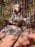 Wooden troll sculpture named Gro sitting cross-legged on a boulder at the Coastal Maine Botanical Gardens, surrounded by tall trees and dappled sunlight.